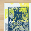 A square art print featuring a stylized navy blue cat surrounded by yellow and white floral patterns, displayed on a light wood background.