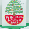 A white greeting card featuring a whimsical illustration of a Christmas tree with cats and the text ’tis the season for cats in trees, shown standing upright on a green envelope against a light gray background.