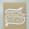 A square kraft paper greeting card with white hand-lettered text reading 'merry Christmas to my person' and small holly motifs, standing upright against a light neutral background with a white envelope partly visible.