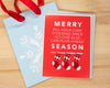A red Christmas greeting card with white text reading 'MERRY FILL-YOUR-OWN STOCKING-SINCE -NO-ONE-ELSE- CAN-PLAN-AHEAD SEASON' and three stocking illustrations, placed on top of a light blue gift bag with a white snowflake and red ribbon on a wood surface.