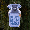Decorative ornament with a raccoon holding a sign saying 'Eat Trash And Be Merry' against a green pine tree background.