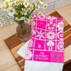 Pink tea towel titled 'The Alphabet in Bloom' with a vase of flowers on a wooden surface
