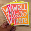 Three colorful greeting cards with 'Well hello there' text held by a hand on a wooden surface.
