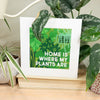 Framed print with 'Home is where my plants are' text surrounded by plants on a wooden surface.