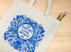 Beige tote bag with blue decorative design and 'Support Local Artists' text on a wooden surface.