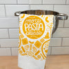 White towel with yellow pasta-themed design and 'More Pasta Please' text hanging over a metal pot on a tiled kitchen wall.