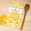 Cotton tea towel with pasta-themed design on a wooden surface with pasta and a wooden spoon.