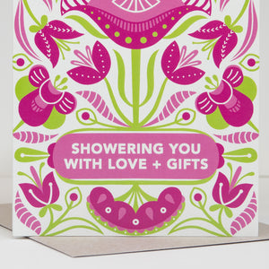 Greeting card with pink and green floral design and text 'Showering you with love + gifts'.