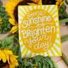 Card with 'Some sunshine to brighten your day' text surrounded by sunflowers on a wooden surface